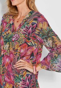 Vibrante blouse met bloemenpatroon in paarse, roze en groene botanische prints. Doorzichtige stof met een V-halslijn en klokmouwen.