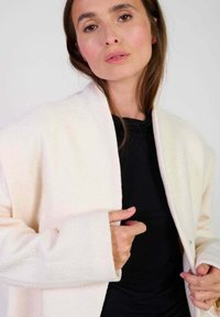 Lichtbeige, gestructureerde jas met een sjaalkraag en een open voorkant. Lange mouwen met brede manchetten en een subtiele glans. Zwarte top eronder.