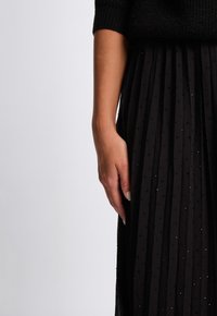 Sort plisseret nederdel med tekstureret mønster, der har små glitrende accenter. Blødt stof, detaljeret med lodrette folder, parret med en sort sweater.