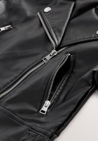 Primer plano de una chaqueta de cuero negra que muestra dos cremalleras plateadas, una cremallera de bolsillo parcialmente abierta y botones a presión de metal en el cuello.