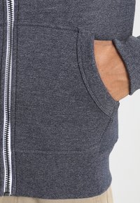 Mano en el bolsillo delantero de una sudadera con cremallera de textura gris oscuro, combinada con pantalones beige.