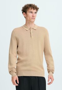 Beige strikket langermet polo-skjorte med en myk tekstur, med krage og tre knappåpninger, pairt med svarte bukser.