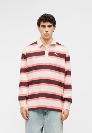 Ung mann med krøllete hår iført en langermet polo med rosa og burgunder stripete mønster og lyseblå jeans, står foran en ensfarget bakgrunn.