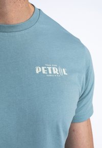 Lichteblauwe katoenen t-shirt met korte mouwen, voorzien van een vervaagd wit logo met de tekst "PETROL HAWAII U.S.A." op de borst. Glad van structuur.