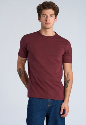 Jonge man met krullend haar, gekleed in een bordeaux T-shirt en blauwe jeans, toont tatoeages op beide onderarmen tegen een effen achtergrond.