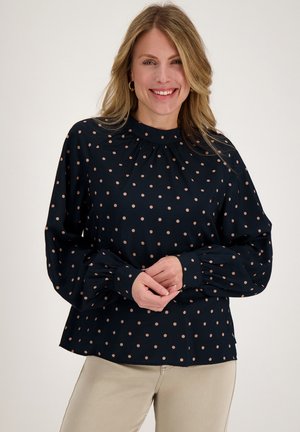 Vrouw die glimlacht, draagt een zwart lang mouwenblouse met beige polka dots en beige broek, staand tegen een egale lichte achtergrond.