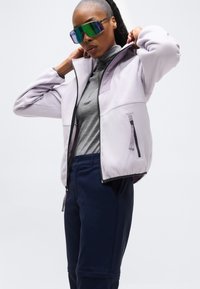 Άτομο που φοράει μεγάλα καθρεφτιστικά γυαλιά ηλίου, ανοιχτό μωβ φλις μπουφάν πάνω από γκρι ζιβάγκο, και μπλε-σκούρα παντελόνια, προσαρμόζοντας το γιακά του μπουφάν.