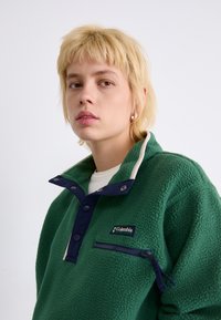 Pull-over en polaire verte avec un col marine et une poche zippée. Comprend des boutons-pression et un patch avec logo. Texture douce et coupe décontractée.