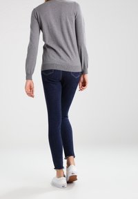Personne marchant au loin vêtue d'un pull gris à manches longues, d'un jean skinny bleu foncé avec des ourlets effilochés, et de baskets blanches.