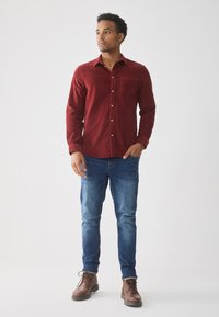 Camisa de pana roja de botones con un bolsillo en el pecho, combinada con unos vaqueros azul denim y botas marrones de cordones. Textura suave con líneas sutiles.