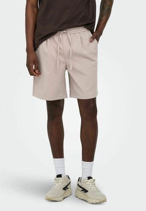 Mann iført beige snorrekortbukser, hvite sokker, beige joggesko og en mørkebrun t-skjorte, står med den ene hånden i lomma.