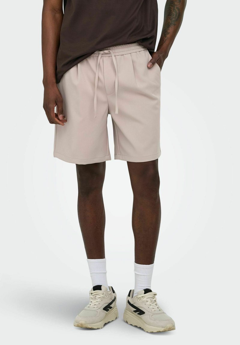 Homme portant un short beige à cordon, des chaussettes blanches, des baskets beiges et un t-shirt marron foncé, debout avec une main dans la poche.