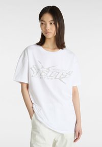 Hvid bomulds t-shirt med en løs pasform, der har et sølvfarvet grafisk logo på forsiden. Korte ærmer og rund halsudskæring.