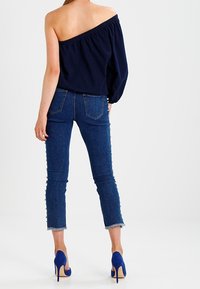 Marinblus utan axlar, i kombination med figurnära blå jeans med sidoknappar. Modellen bär blå klackar och visar en avslappnad, trendig stil.