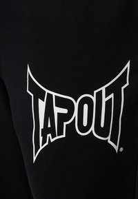 Black sweatpants featuring a large white logo that reads "TAPOUT." The fabric appears smooth with a soft texture.