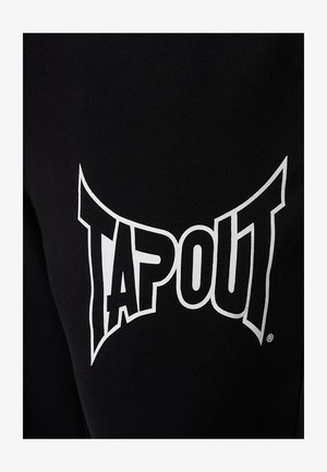 Melnas sporta bikses ar lielu baltu logotipu, kas saka "TAPOUT". Audums izskatās gluds ar mīkstu tekstūru.