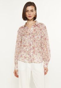 Bloemige zijden blouse met lange mouwen, knoopsluiting en paillettenaccenten. Lichtroze achtergrond met kleine paarse en gele bloemenpatronen.