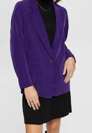 Persona che indossa un blazer viola su un vestito nero, con anelli d'argento alle dita e una collana a catena d'argento visibile.