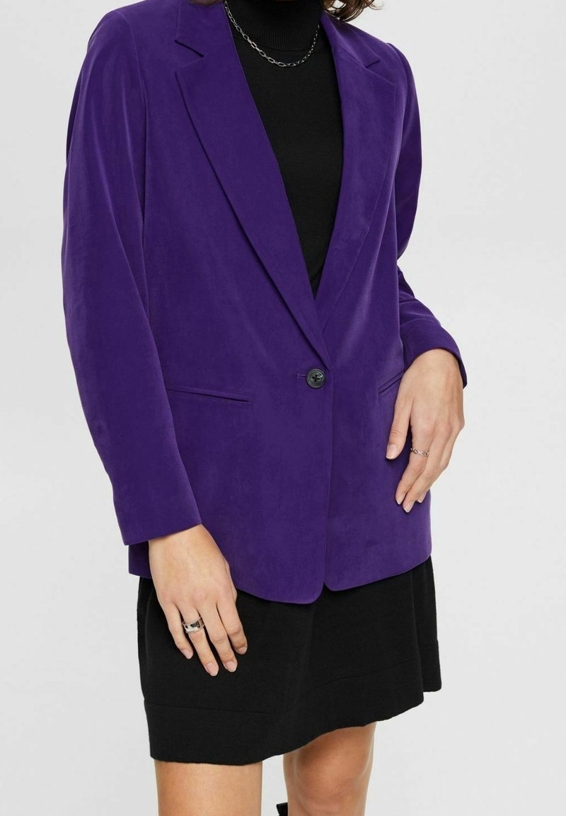 Personne portant un blazer violet sur une robe noire, avec des bagues argentées aux doigts et un collier chaîne argenté visible.