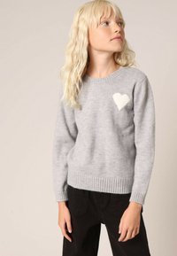Grijze gebreide trui met een geribde zoom, voorzien van een wit hartvormig ontwerp op de linkerkant van de borst. Gecombineerd met zwarte broek.