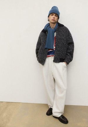 Jachetă texturată neagră și gri, căciulă tricotată albastră, eșarfă albastră, cămașă cu dungi și pantaloni de trening gri deschis, asortați cu încălțăminte întunecată.