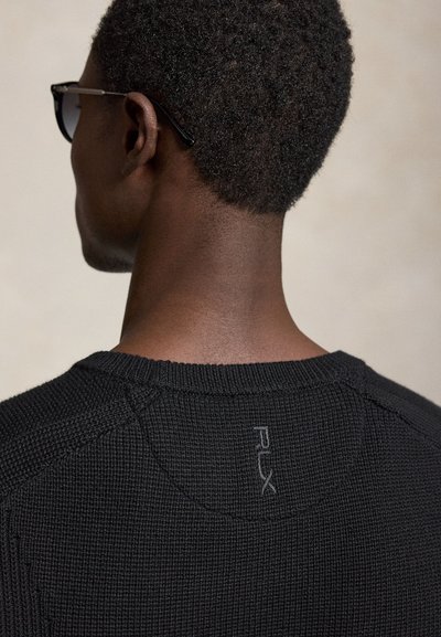 Homme aux cheveux courts et bouclés portant des lunettes de soleil noires et un pull texturé noir avec un petit logo PLX à l'arrière du col.