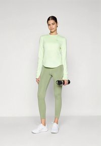 Kvinne står i lys grønn langermet topp, matchende leggings, hvite sneakers, holder en svart vannflaske mot en enkel bakgrunn.