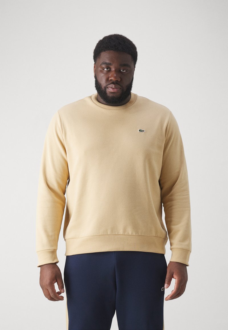 Beige Sweatshirt aus Baumwolle mit Rundhalsausschnitt, gerippten Bündchen und einem kleinen grünen Logo auf der Brust, kombiniert mit dunkelblauen Hosen.