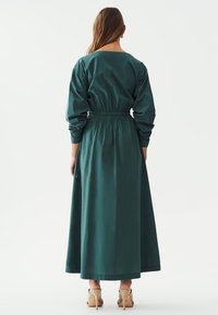 Pitkä turkoosi mekko, jossa on pitkät hihat, joustava vyötärö ja virtaava siluetti, valmistettu sileästä kankaasta, esitelty takaa.