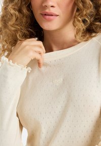 Close-up van een vrouw met krullend haar die een off-white, textuurverzorgd shirt met lange mouwen draagt, versierd met een klein hartjespatroon en gerimpelde mouwen.