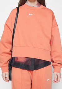 Överdimensionerad orange sweatshirt med långa ärmar, med en vit Nike-logotyp på bröstet och en lager av svart skjorta som kiknar fram under.