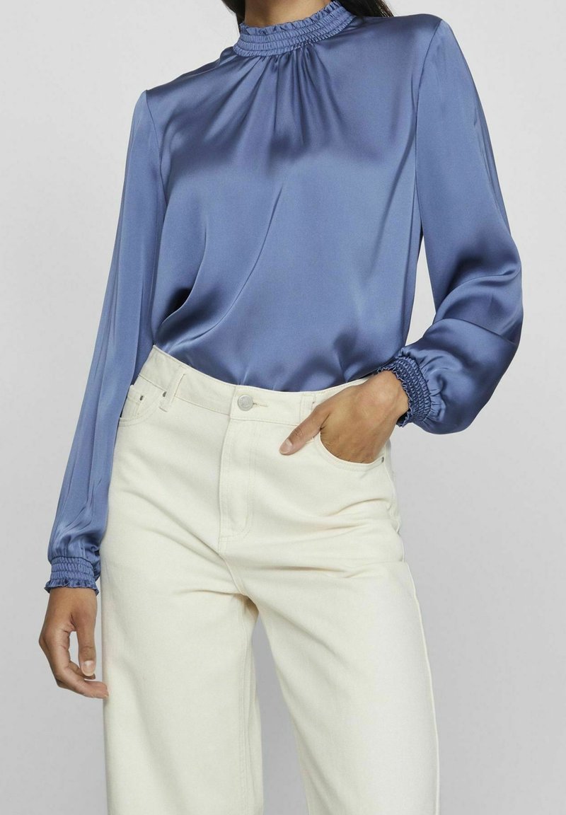 Blaue Satinbluse mit gerafftem Halsausschnitt und Manschetten, kombiniert mit hochgeschnittenen, cremefarbenen Hosen mit glatter Textur und weitem Bein.