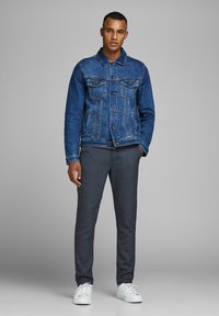 Giacca di jeans blu scuro con taglio classico, tasche sul petto e cuciture a contrasto. Abbinata a pantaloni neri affusolati e sneakers bianche.