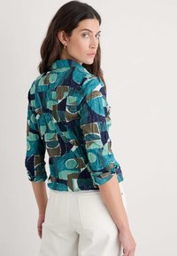 Skjorte med krave i en krøllet tekstur, prydet med et flerfarvet geometrisk mønster i teal, marineblå, grøn og brun. Lange ærmer opprullet.