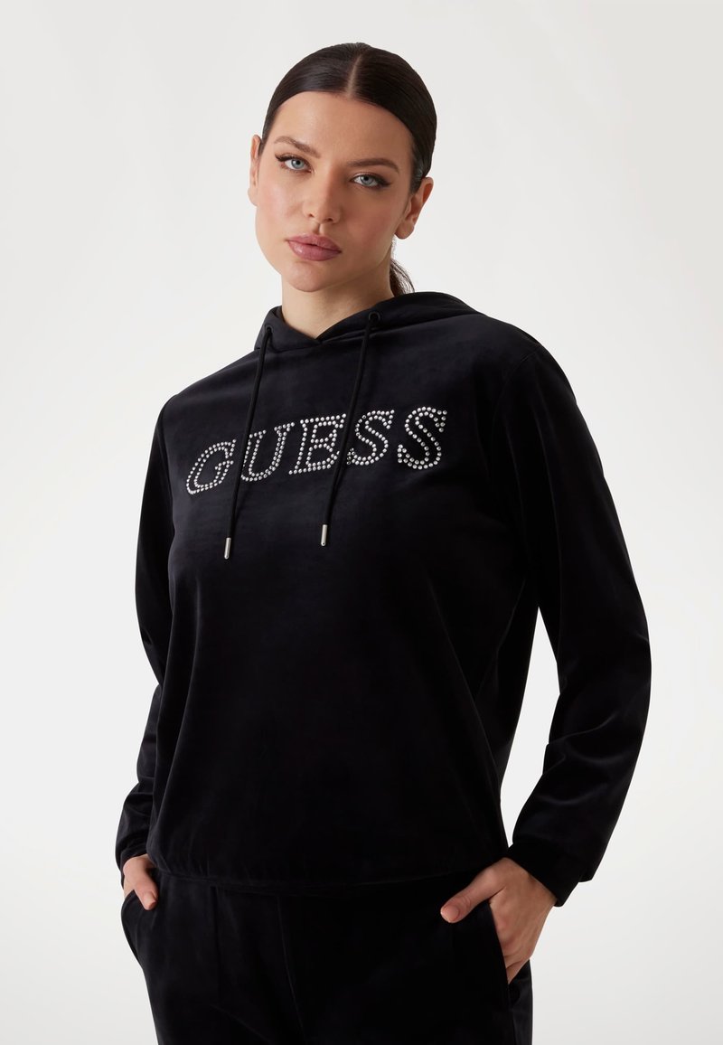 Čierna mikina s kapucňou vyrobená z jemnej tkaniny, s nápisom "GUESS" osadeným kryštálmi. Obsahuje predné vrecko a nastaviteľnú kapucňu so šnúrkou.