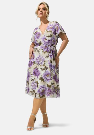 Femme portant une robe portefeuille mi-longue à imprimé floral violet, manches courtes volantées et talons ouverts argentés sur un fond uni.