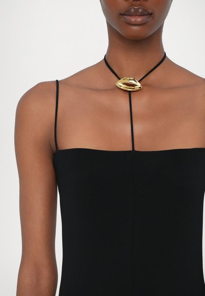 Μαύρο αμάνικο ένδυμα με ίσια λαιμόκοψη που διαθέτει ένα χρυσό μεταλλικό κρεμαστό κόσμημα σε μαύρο κορδόνι, αναδεικνύοντας την απαλότητα και τη μορφή του.