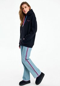 Naine seisab musta kapuutsiga, millel on roosad ja sinised paelad, helesiniste pükstega, millel on roosad triibud, ja mustade libisevate kingadega.