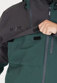 Veste imperméable verte et grise avec une poche avant à fermeture Velcro, logo de la marque "SLOPE", et un plastron zippé.