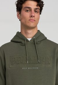Felpa verde oliva con tasca a marsupio, caratterizzata da un testo in rilievo "B.R.E.A.K.E.R!" e "SELF BELIEVER" in bianco sottostante. Con corde regolabili.