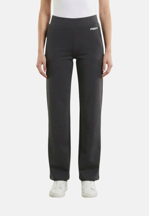 Persona che indossa pantaloni neri a gamba dritta con logo vicino alla vita, scarpe da ginnastica bianche e una maglietta bianca, in piedi su uno sfondo chiaro uniforme.