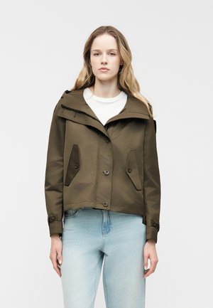 Femme aux longs cheveux blonds portant une veste boutonnée vert olive avec des poches à rabat sur un haut blanc et un jean bleu clair, face à l'avant.