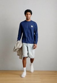 Μπλούζα με μακριά μανίκια σε ναυτικό μπλε με γραφικό φοίνικα, ανοιχτόχρωμο τζιν σορτς, λευκά αθλητικά παπούτσια και μια μπεζ μπουφάν περασμένη στον βραχίονα. Φόντο ξύλινο πάτωμα.