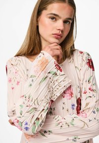 Svijetloružičasta bluzica s nabranim detaljima i cvjetnim uzorcima u raznim bojama; ima široke, nabranne rukave i rebrasti tekstur.