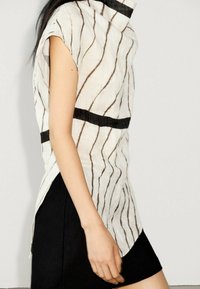 Haut blanc sans manches avec un motif rayé noir et un col montant, associé à une jupe noire. Le tissu semble texturé et léger.