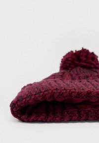 Červená pletená čiapka s textúrovaným vzorom a vrchovým pom-pomom. Materiál vyzerá jemne a hrubo s tmavšími škvrnami.