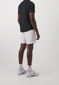 Man som bär svart t-shirt, vita shorts, vita Nike-sneakers och vita strumpor, står mot en ljus enfärgad bakgrund, sedd från baksidan.