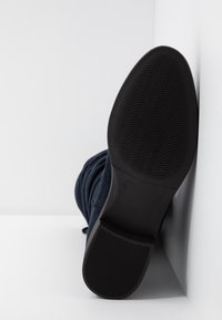 Semelle d'une botte noire avec un profil texturé, inclinée contre un mur d'angle blanc, indiquant la taille 37 près de la voûte plantaire.