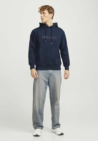 Mørkeblå pullover hoodie med frontlomme, snoretræk i hætten og teksten "ORIGINALS" trykt i midten. Båret sammen med lyse jeans.