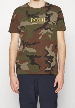 Mann trägt ein grün-braunes Camouflage-T-Shirt mit gelbem "POLO Ralph Lauren"-Text und dunkelblauer Hose, steht vor weißem Hintergrund.